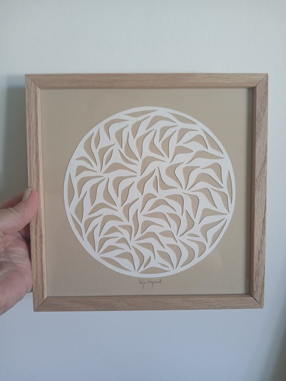 Hånd holder firkantet ramme med abstrakt cirkulært mønster i hvidt papir mod beige baggrund.