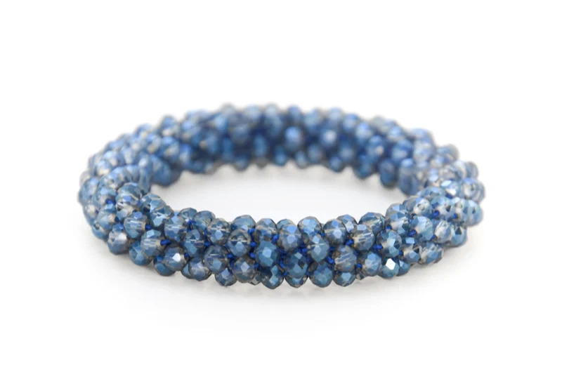 Blåt armbånd lavet af facetterede perler snoet i et tæt, rundt mønster på en hvid baggrund.