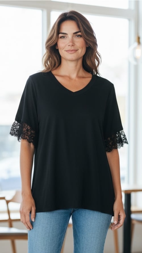 Kvinde iført sort T-shirt med blonder på ærmerne og blå jeans, stående indendørs foran vindue. (1)