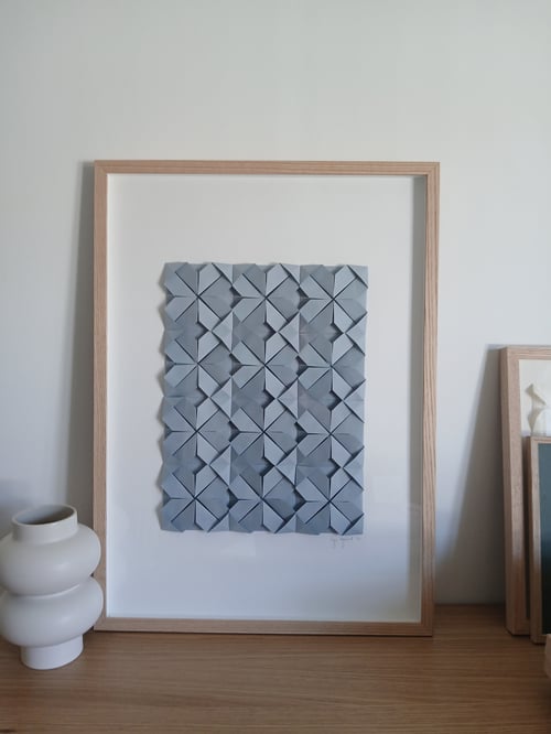 Indrammet kunstværk med tredimensionelle, foldede grå firkanter arrangeret i et mønster, placeret på en træoverflade ved siden af en hvid, moderne keramikvase. (2)