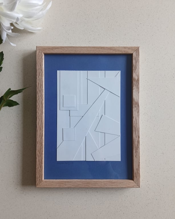 Minimalistisk abstrakt kunstværk med hvide geometriske former arrangeret på en blå baggrund i en træramme, placeret på en lysegrå bordplade med en hvid blomst i øverste venstre hjørne.