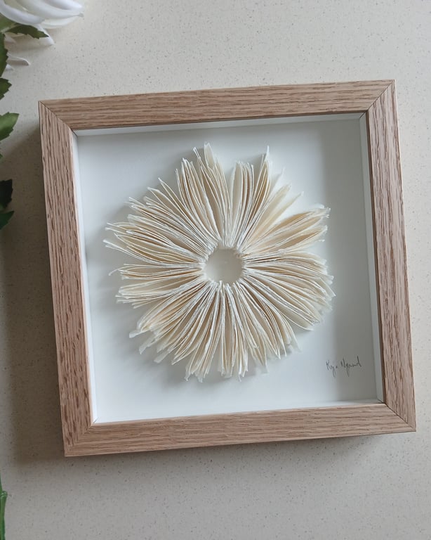Billedramme med kunstværk bestående af foldede papirblade arrangeret i en cirkel med et hul i midten.