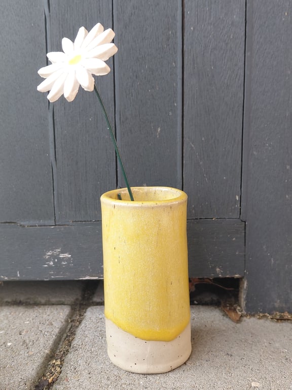 En gul og beige keramisk vase med en enkelt kunstig hvid blomst placeret foran en mørkegrå trædør på en flisebelagt overflade.