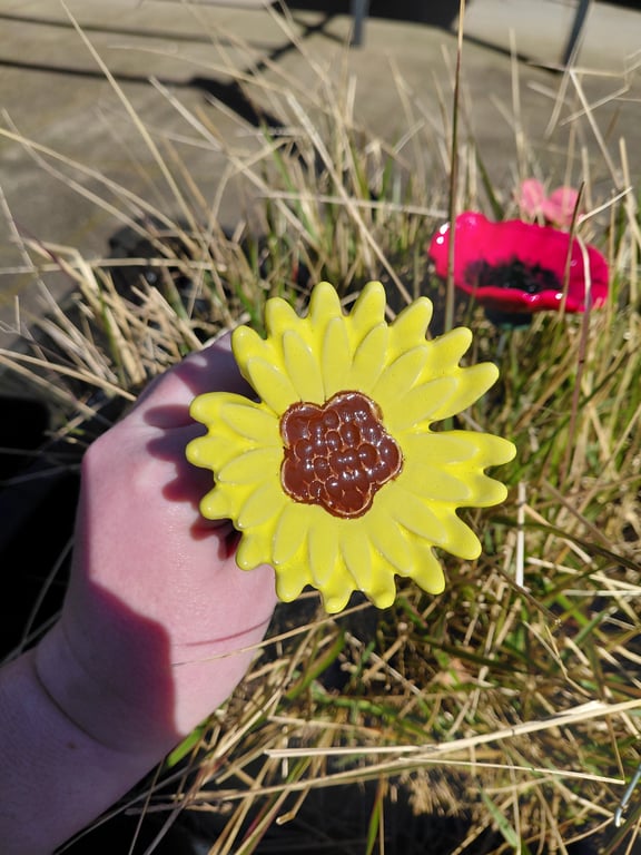 En hånd holder en keramisk gul solsikke med brunt midtpunkt over tør græs med en rød keramisk blomst i baggrunden.