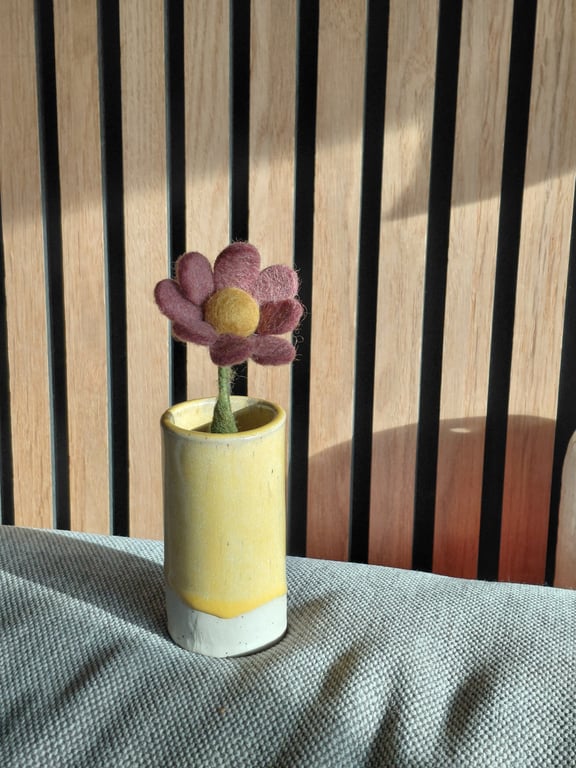 En blomst lavet af filt med lilla blomsterblade og en gul midte i en lille gul og hvid keramikvase, placeret foran en træbeklædt væg.