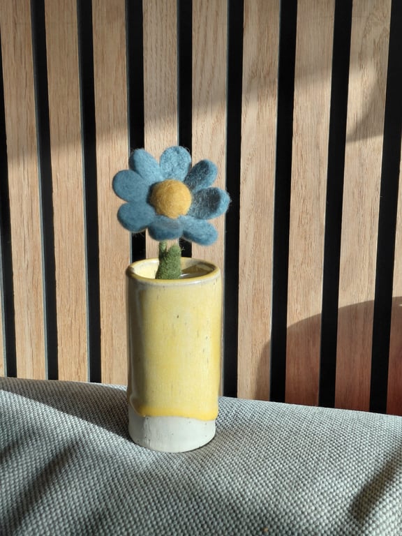 Blå filtblomst med gul midte i lille gul keramikkrukke foran træpaneler, stående på grå stofoverflade.