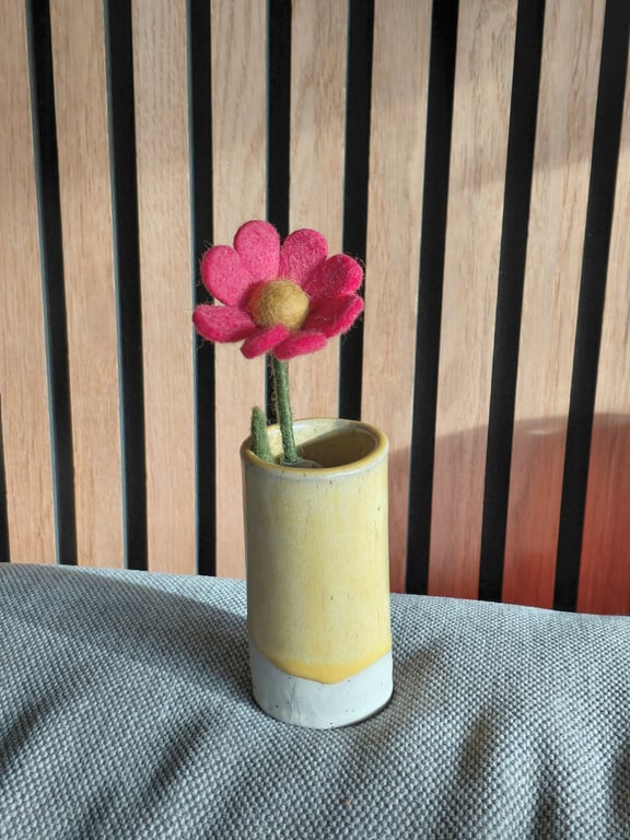 En gul og hvid keramisk vase med en rød filtblomst og grøn stilk, placeret på en grå pude foran en trævæg med sorte lodrette striber.