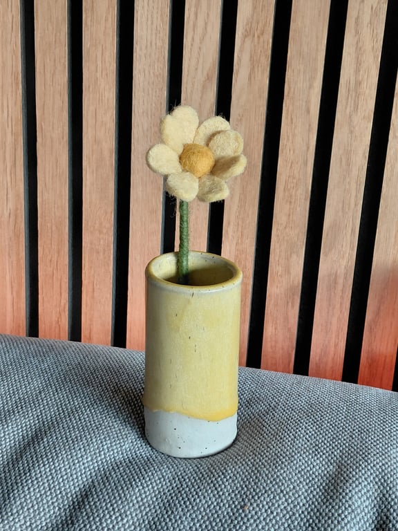 En filtblomst med gule kronblade i en lille gul-hvid keramisk vase, placeret på en grå stofoverflade foran en trævægliste.