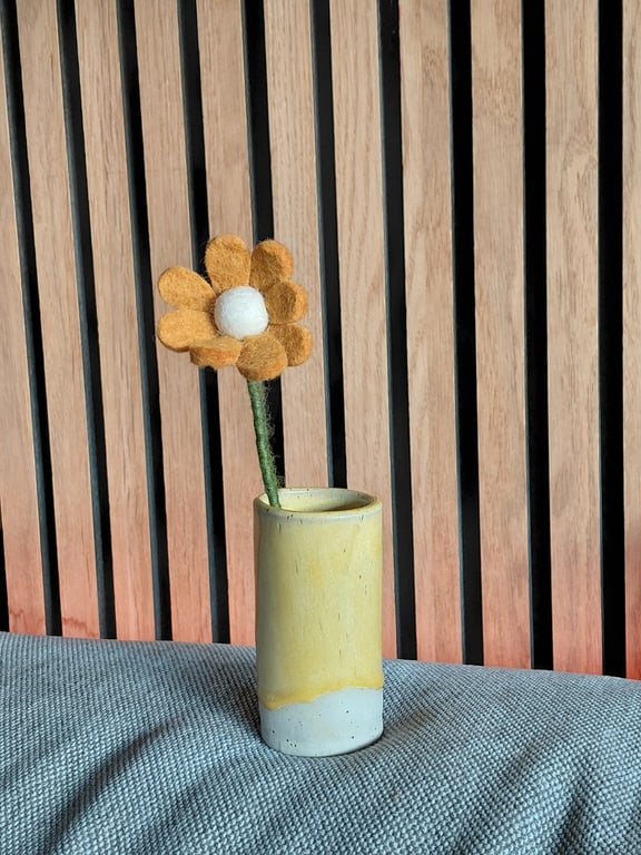 En gul og hvid keramikvase med en filtblomst placeret på en grå stofoverflade foran en væg med vertikale træpaneler.