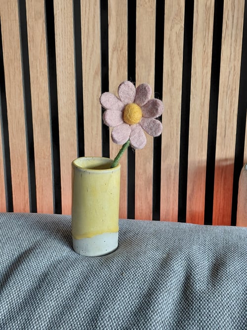En lille gul og hvid keramikvase med en filtblomst, placeret på en grå tekstileret overflade mod en baggrund af lodrette træpaneler. (1)