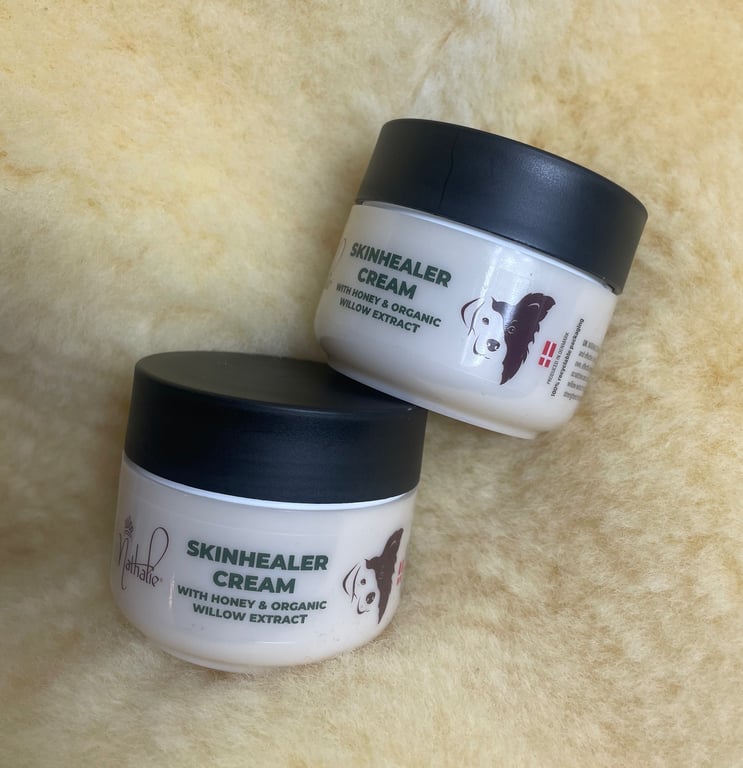 To behold two small white cream jars with black lids labeled "Skinhealer Cream with Honey & Organic Willow Extract" placed on a soft beige surface.