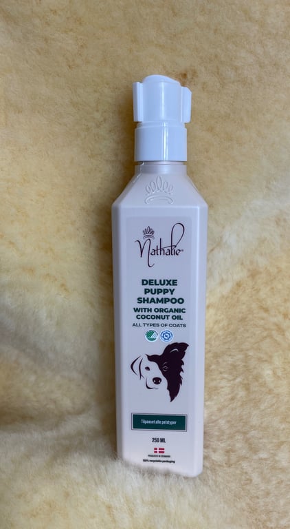 Flaske med Nathalie Deluxe Puppy Shampoo med økologisk kokosolie til alle pels typer, placeret på blødt lyst stof.