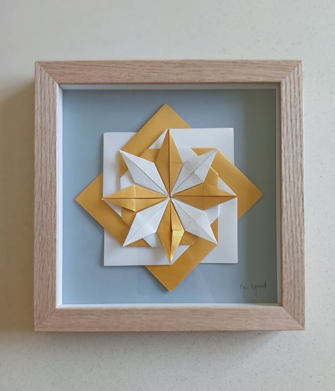 Origami-stjerne i hvidt og guldpapir, monteret i en lysebrun træramme med lyseblå baggrund.