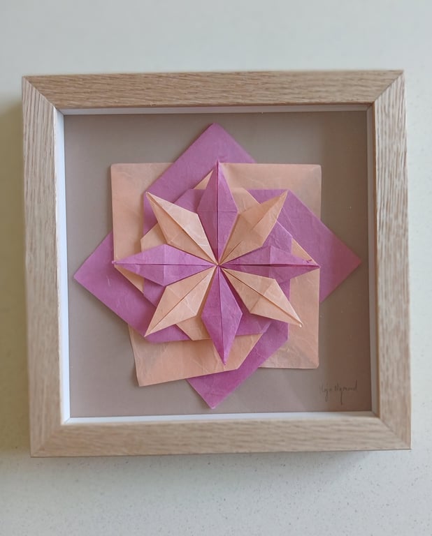 Origamiskulptur i lyserøde og beige nuancer formet som en geometrisk blomst, indrammet i en lys træramme.