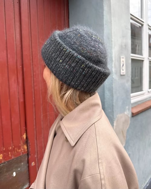 Kvinde set bagfra iført beige frakke og grå strikket hue, stående foran rød dør og grå bygning med vindue. (1)