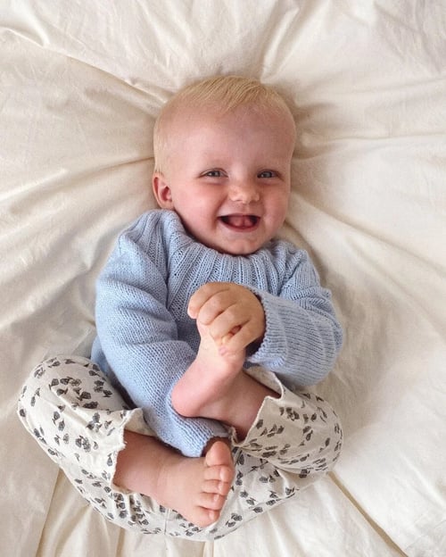 Smilende baby iført lyseblå striktrøje og mønstrede bukser, der ligger på hvidt sengetøj og holder sin fod. (1)