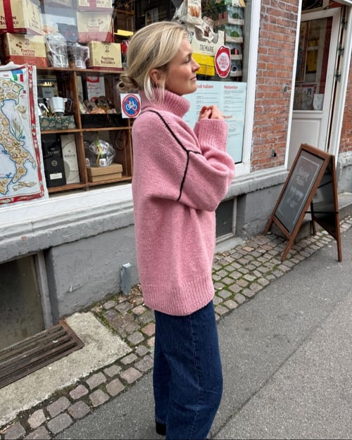 En kvinde iført en lyserød striktrøje står på en brostensbelagt fortov foran en butik med glasvindue fyldt med diverse varer. (1)