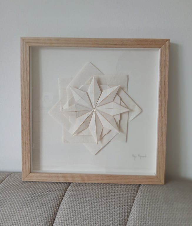 Indrammet papirkunst med et geometrisk, foldet stjernemønster i hvidt papir, placeret på en lys væg og en grå sofa.
