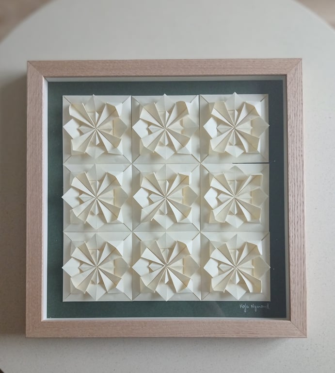 Indrammet kunstværk bestående af ni hvide, tredimensionelle papirblomster arrangeret i et 3x3 grid på en mørkegrøn baggrund.