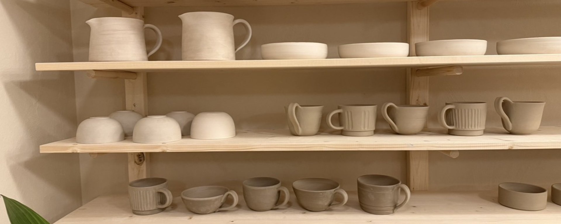 Hylder med forskellige umalede lerkrus, kander, skåle og tallerkener i et keramisk værksted.