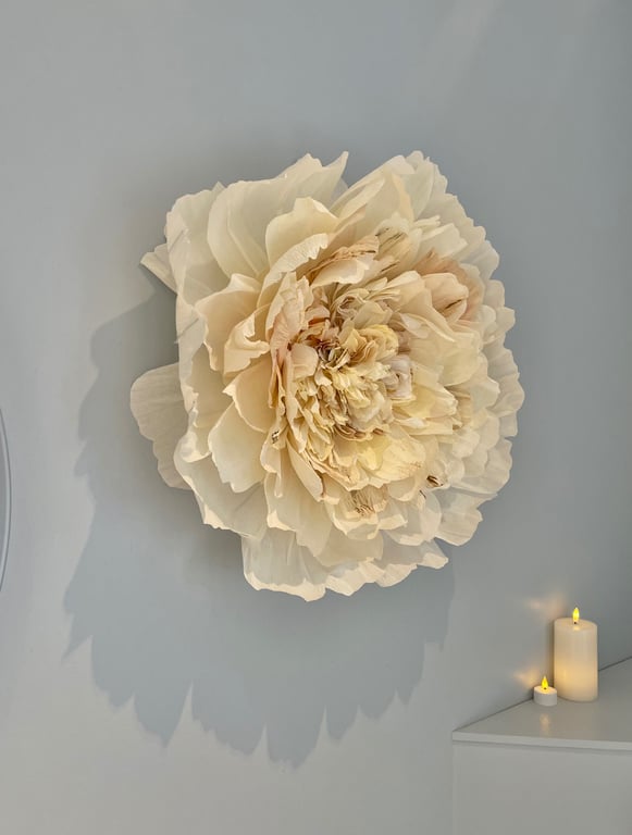 Stor dekorativ blomst i lyse beige nuancer hængende på en grå væg med to tændte lys på en hvid overflade ved siden af.