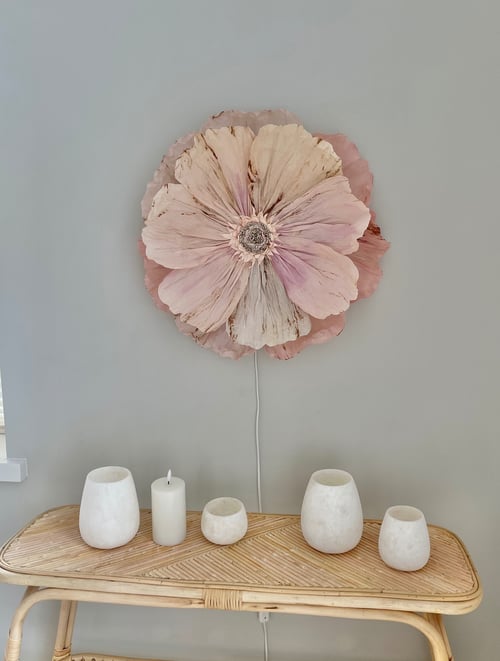 Stor, lyserød blomsterdekoration hænger på en grå væg over et bord med fire hvide lysestager og et tændt stearinlys. (1)