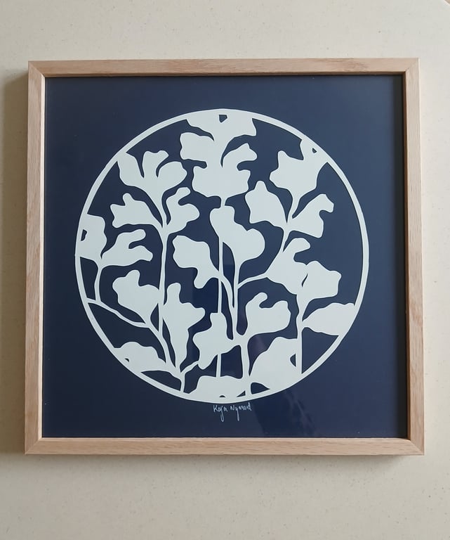 Billedet viser en firkantet træramme med en mørk baggrund og et cirkulært, hvidt, abstrakt bladmotiv i midten.