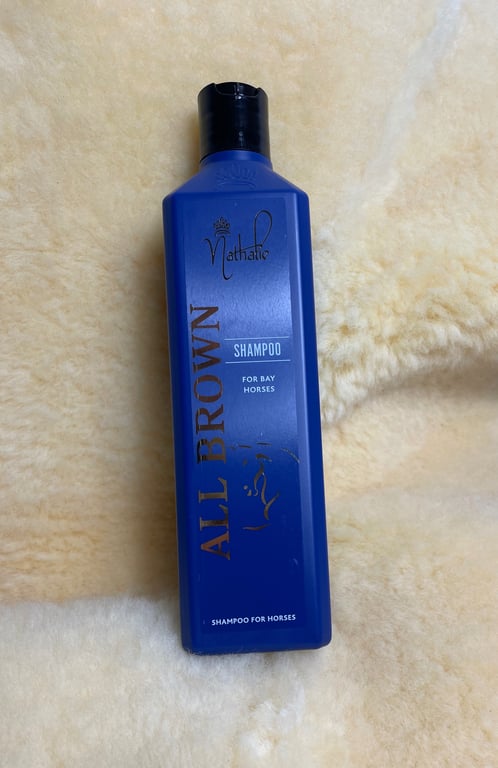 Flaske med blå shampoo til brune heste, mærket "Nathalie All Brown Shampoo for Bay Horses" på en lyst uldagtigt underlag.