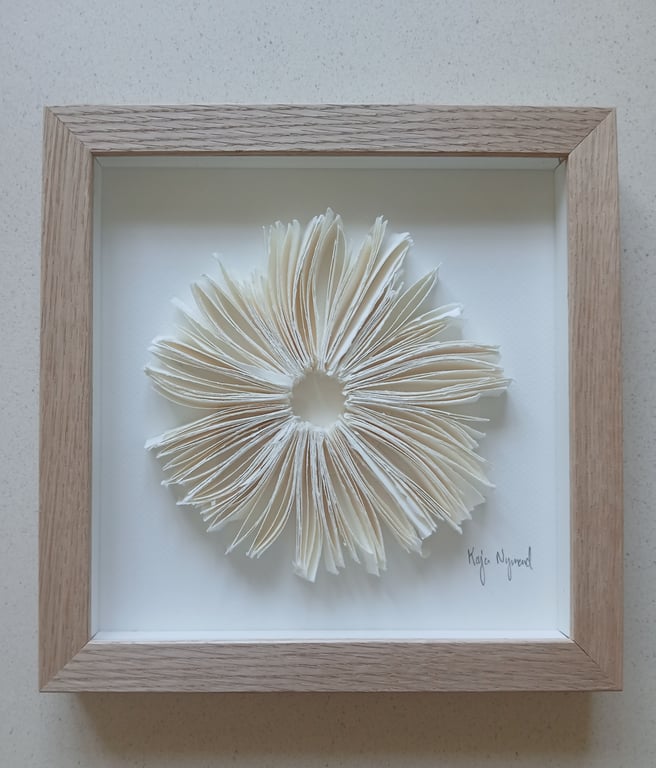 Indrammet kunstværk med foldede papirark arrangeret i en rund blomstlignende form på hvid baggrund.