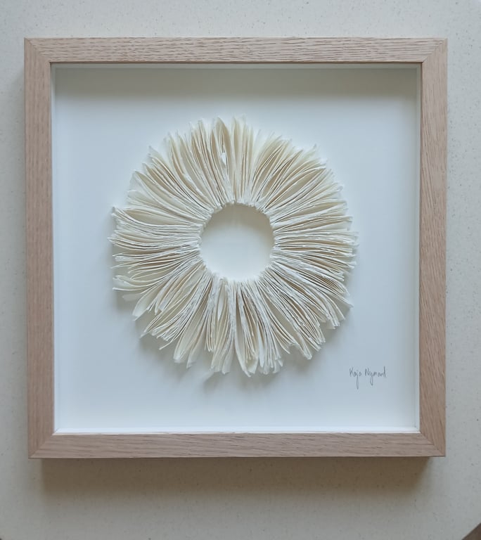 Indrammet kunstværk med en cirkel af udslåede, foldede hvide papirark, der danner en blomstlignende form på hvid baggrund.