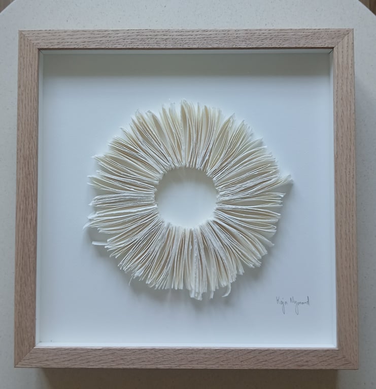 Indrammet kunstværk med en cirkulær form lavet af foldede hvide papirsider, der danner en blomst-lignende struktur på en hvid baggrund.