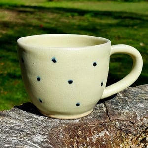 Kop med hank i mat gul glasur med sorte prikker.   – produktbillede