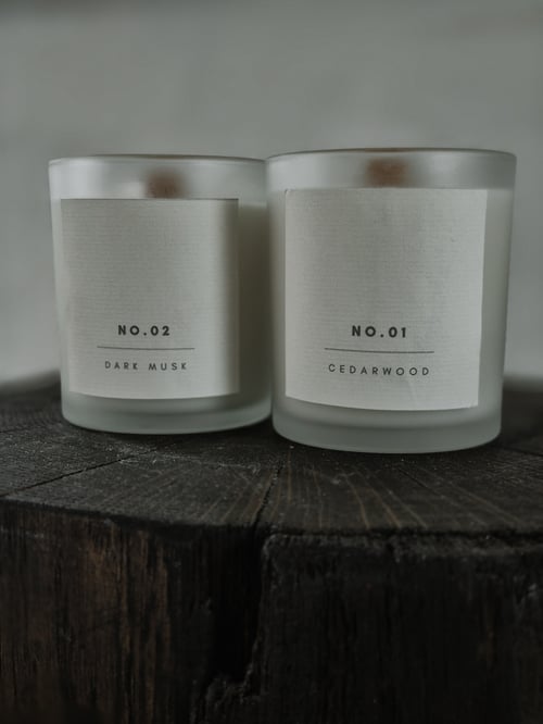 To duftlys i matteret glas med mærkater, der angiver duftene "No.02 Dark Musk" og "No.01 Cedarwood," placeret på en rustik træoverflade. (1)