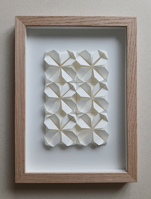 Indrammet papirkunst med en tredimensionel geometrisk mønster bestående af hvide foldede papirformer arrangeret i fire rækker og tre kolonner.