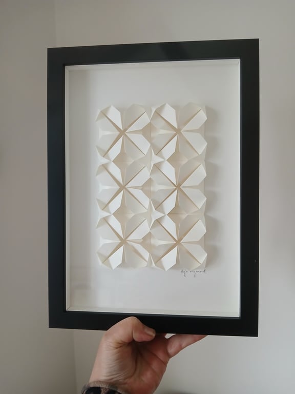Hånd, der holder en sort indrammet 3D-kunst med geometriske hvide papirformer arrangeret i et mønster på en hvid baggrund.