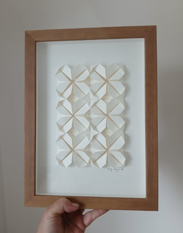 Et håndlavet hvidt papirkunstværk med geometriske foldede mønstre i en brun træramme, holdt i hånden mod en neutral baggrund.