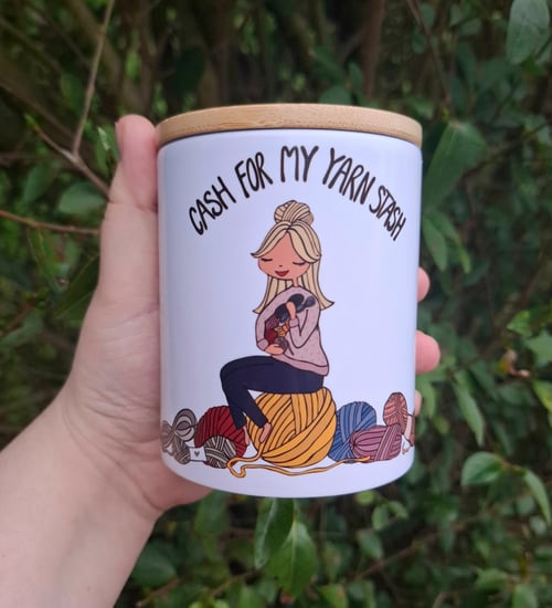 Hånd, der holder en hvid krukke med låg, dekoreret med en tegning af en kvinde omgivet af garnnøgler og teksten "Cash for my yarn stash". (1)