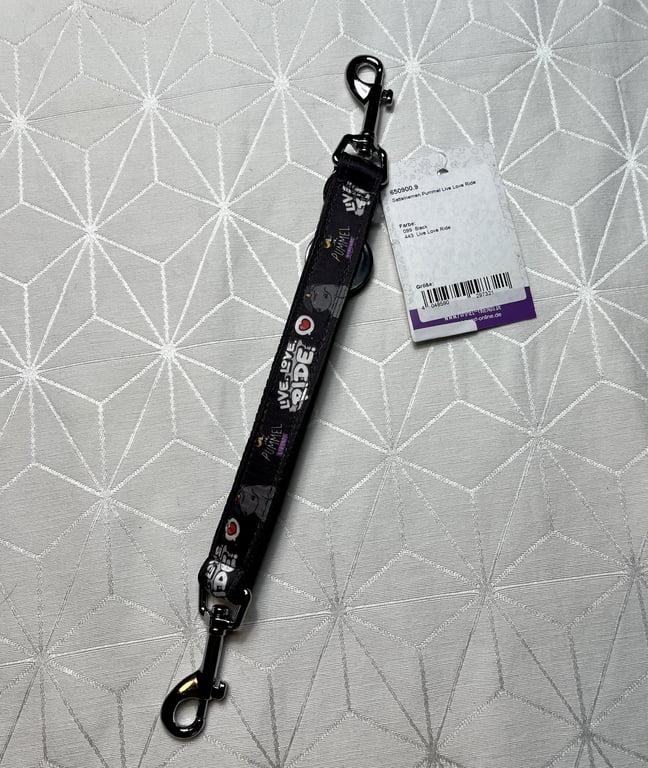 Sort hundesnor med hvide og lilla tekster og motiver samt metalclips i begge ender, liggende på hvidt mønstret stof.