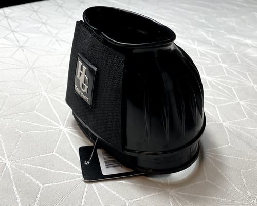 Sort gummibeskytter til hove med elastisk bånd og HG-logo placeret på et lyst bord med geometrisk mønster. (1)