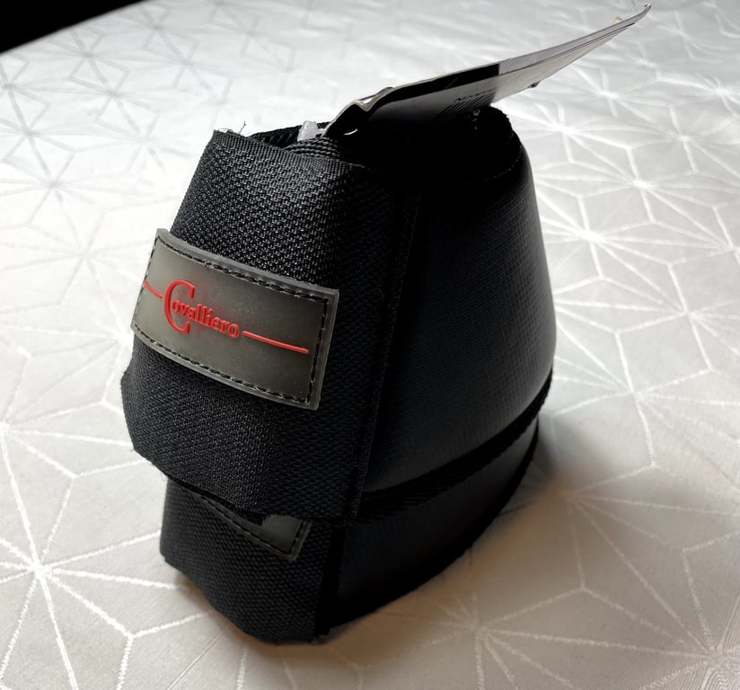 Sort beskyttelsesbandage til hesteben med mærket "Covalliero" i rød skrift på sort baggrund, placeret på en lysegrå bordduk med geometrisk mønster.