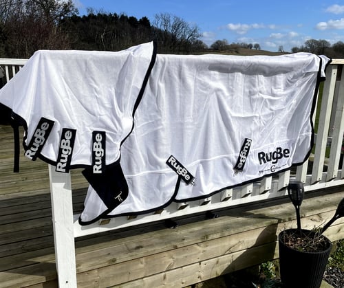 Hvidt tæppe eller dækken med sort kant og flere mærker med teksten "RugBe" hængende over et hvidt gelænder på en træterrasse udendørs. (1)