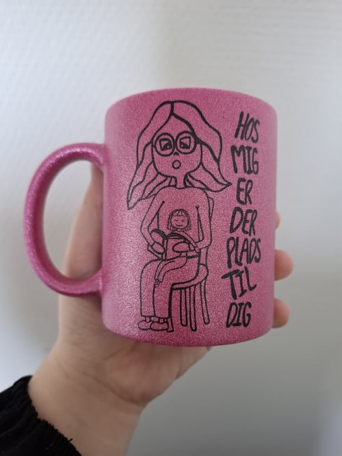 Hånd, der holder en glitrende pink krus med en tegning af en kvinde, der ammer et barn, og teksten "Hos mig er der plads til dig". (1)