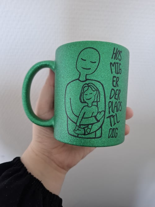 En hånd holder en grøn glitrende krus med en tegning af to personer, hvor den ene holder den anden, og teksten "Hos mig er der plads til dig" skrevet ved siden af. (1)