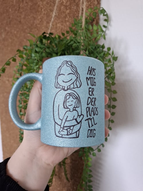 Hånd, der holder en glitrende blå krus med tegning af to omfavnende figurer og dansk tekst "Hos mig er der plads til dig" foran grøn plante og korkvæg. (1)