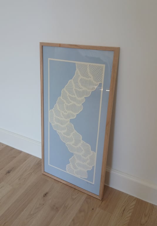 Rektangulært kunsttryk med bølgende hvide linjer på lyseblå baggrund, indrammet i lyst træ og placeret op ad en hvid væg på lyst trægulv.