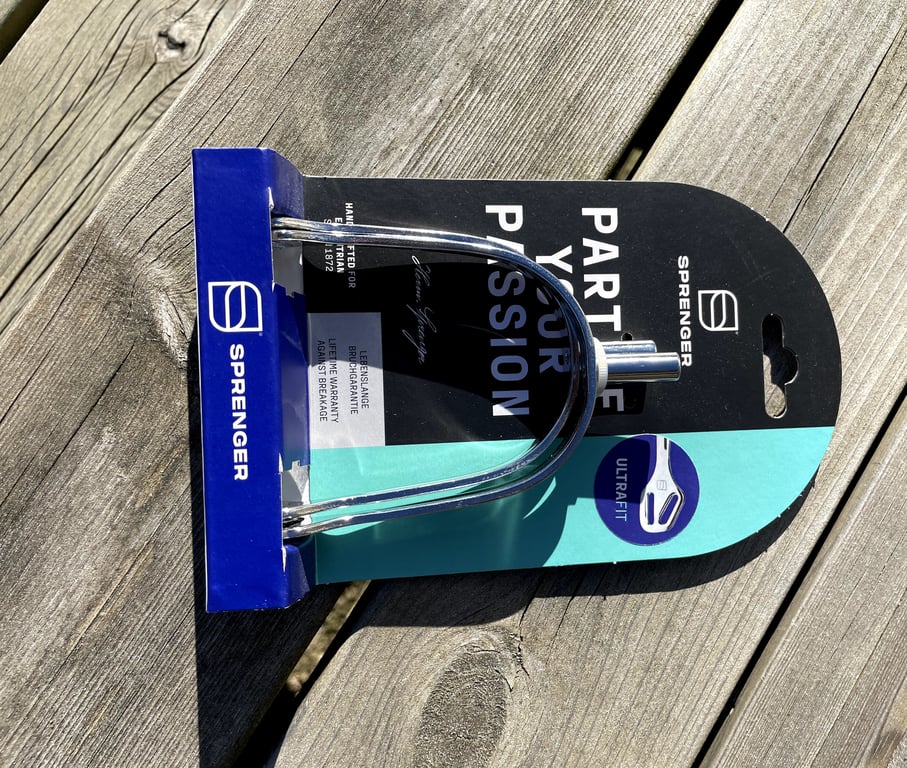 Sprenger ultrafit stigbøjle i metal på sort og turkis emballage, placeret på træflade.