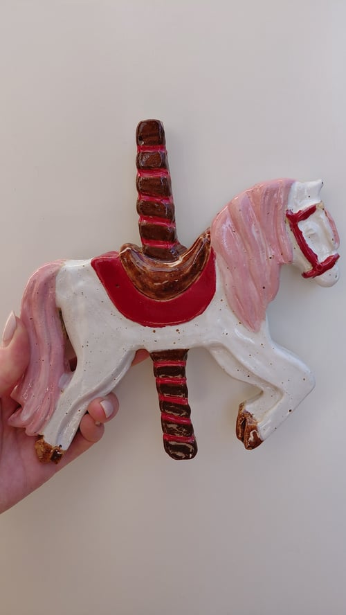 Hånd, der holder en keramisk karrusel-hest med lyserød man og hale, rød sele og hestestang. (1)
