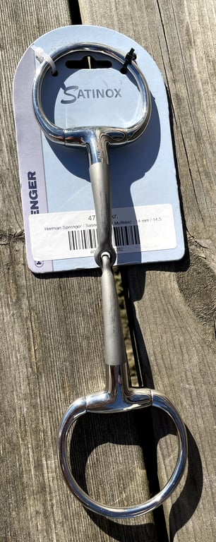 Billede af et metalbid med to ovale ringe, placeret på en træflade med en papæske med teksten "Satinox".
