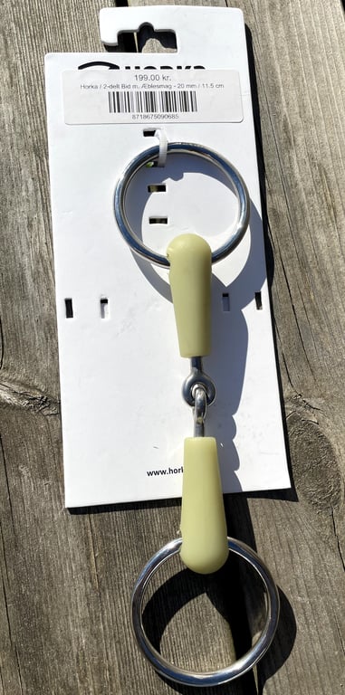 Bidsel med gummibelægning og metalringe, vist på hvidt ophængskort med pris og produktinformation, lagt på træoverflade.