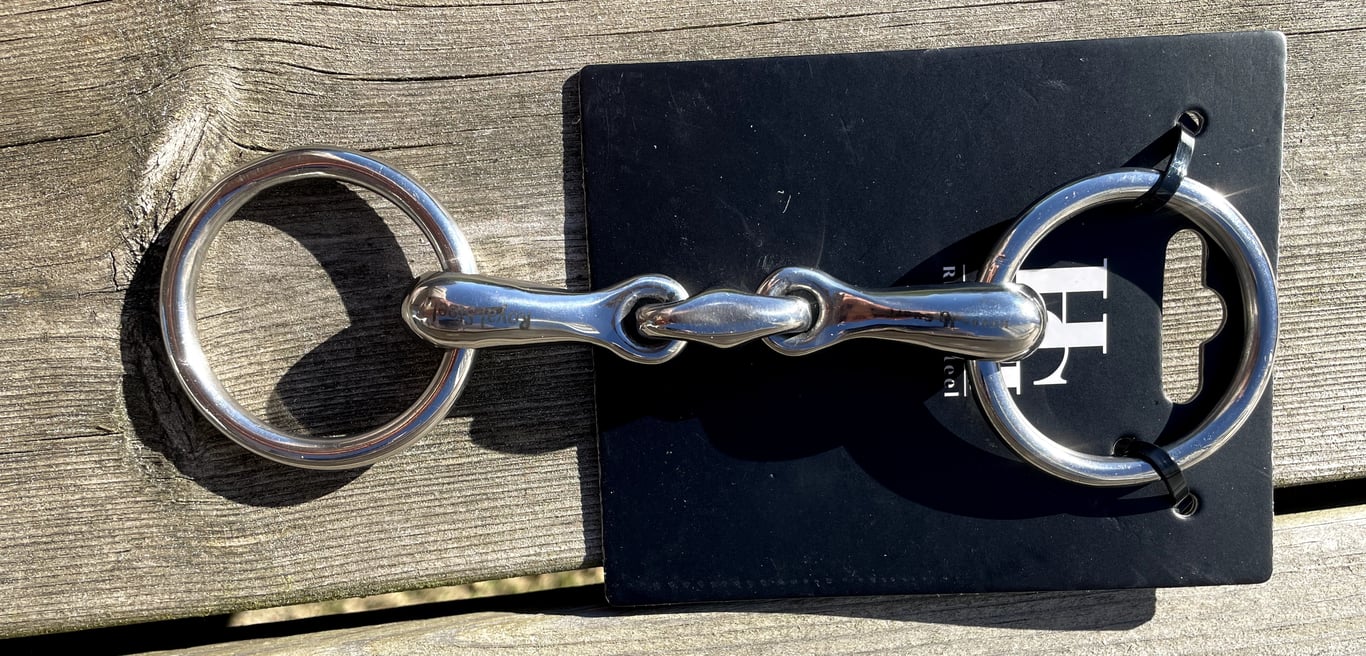 Bidsel med to store metalringe og et ledet mellemliggende stykke, lagt på en sort baggrund og et træbord i solen.